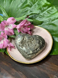 pyrite heart on a pink plate close up with pink flower with green leaf on a wood table