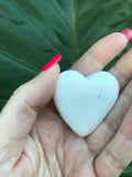 Heart-shaped stone held in a hand with pink nail polish against a green leafy background