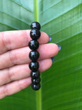 a black bead shungite bracelet being held up by a woman's hand against a dark green leaf