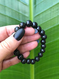 a black bead shungite crystal bracelet being held by a woman with black fingernail polish against a green white bird of paradise leaf
