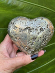 a large pyrite heart being held by a woman's hand in front of a green leaf