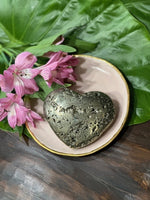 pyrite heart on a pink dish by pink flowers sitting on a dark green leaf on a wood table