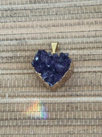 a raw heart shaped amethyst that is deep purple with gold edge pendant on a tan natural fiber background