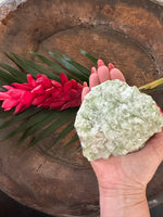 a light Green Apophyllite Crystal being held in a woman's hand over a bowl with a red ginger flower and green leaf