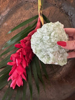 a light Green Apophyllite Crystal being held by a woman with red nails over a red ginger flower and green leaf in a wooden bowl