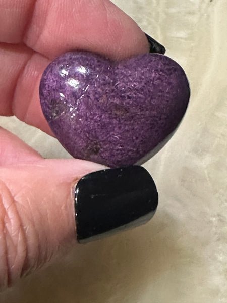 one Purpurite crystal heart being held close up between a woman's fingers with black nail polish