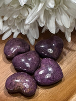 several Purpurite crystal hearts that have a gold sheen by a white flower on a wooden tray