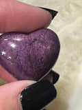 a Purpurite crystal heart close up with a golden sheen being held by a woman between her fingers