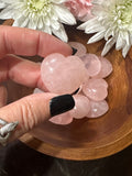 small pink rose quartz crystal heart held by a woman's fingers over a bowl of small rose quartz hearts in a bowl by white and pink flowers