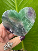 A rainbow fluorite heart shaped crystal dish being held up by a woman's hand against a green leaf