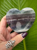 a rainbow fluorite heart shaped crystal dish that has bands of purple and green across held by a woman's hand in front of a green leaf