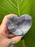 a purple and green band that goes more to the right rainbow fluorite heart shaped crystal dish held up in between a woman's hand in front of a green leaf