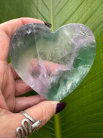 a light purple and mostly green rainbow fluorite heart shaped crystal dish held between a woman's fingers against a leaf background