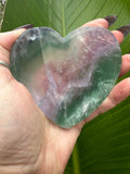 a light purple with dark green bands across the top right rainbow fluorite heart shaped crystal dish held in the middle of a woman's hand against a dark green leaf