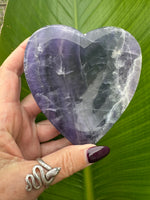 a dark purple and green bands going up and down rainbow fluorite heart shaped crystal dish held up by a woman's hand in front of a dark green leaf