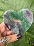 a  dark green and purple band that goes across and down the bottom right rainbow fluorite heart shaped crystal dish held between a woman's fingers against a dark green leaf