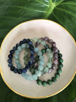 a set of crystal bracelets including sodalite aquamarine prehnite lepidolite ruby zoisite crystal bracelets on a pink plate with gold rim on a green leaf