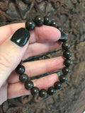 brown bead bronzite crystal bracelet being held by a woman with black nails againsta a dark indian carved wood piece