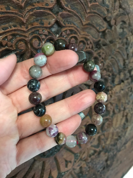 mixed tourmaline crystal bead bracelet - black purple yellow white gray green beads - held by woman's hand against wooden carved board