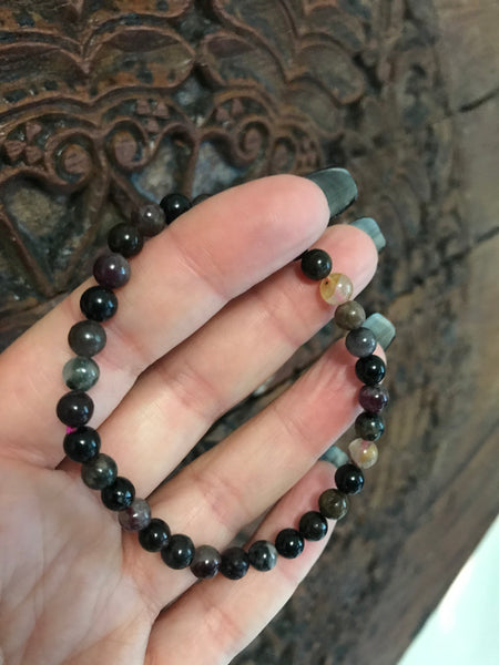 a mixed color - purple black green brown and yellow mixed tourmaline bracelet being held by a woman's hand against a carved indian wood board