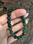 a dark green with lighter green hues bead moss agate crystal bracelet being held by a woman's hand on a carved indian wood board