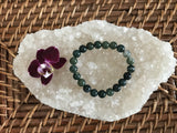 a dark green and lighter hue green moss agate crystal bead bracelet near a maroon orchid flower on a white apophyllite crystal on a wicker tray