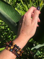 three tiger's eye multi-color crystal bead stretch bracelet on a woman's hand in front of green leaves