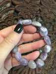 a light purple gray and white tumbled amethyst stretch bracelet being held by a woman's against a dark carved wooden indian board