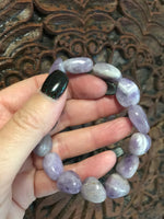 a light purple gray and white tumbled amethyst stretch bracelet being held by a woman's against a dark carved wooden indian board