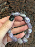 a light purple gray and white tumbled amethyst stretch bracelet being held by a woman's against a dark carved wooden indian board