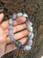 a light purple gray and white tumbled bead amethyst crystal bracelet being held up by a woman's hand against a wooden indian carved board