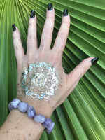 a light purple gray and white tumbled bead amethyst crystal bracelet on a woman's hand with a gold tattoo with black nails against a green palm leaf