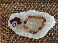 an orange tumbled carnelian crystal  bracelet by a maroon orchid on a white apophyllite crystal on a wicker tray