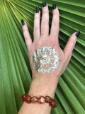 an orange tumbled carnelian crystal bracelet on a woman's hand with a gold tattoo and black nails against a green palm leaf