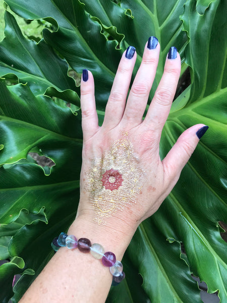 a tumbled fluorite crystal bracelet on a woman's wrist with a gold tattoo and blue nails against a green leaf