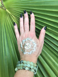 3 light green tumbled prehnite crystal stretch bracelets on a woman's arm with a gold tattoo with black fingernails against a green palm leaf