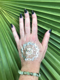 a light green tumbled prehnite crystal stretch bracelet on a woman's arm with a gold tattoo and black nails against a green palm leaf